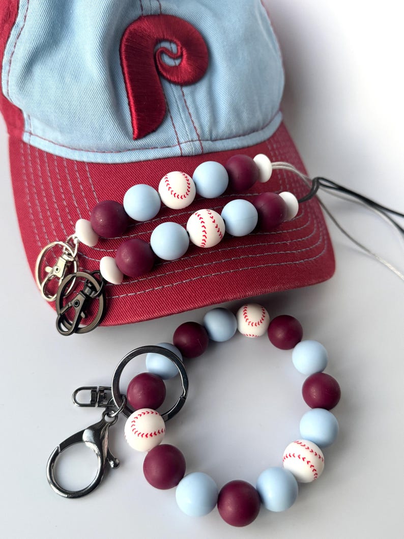 May include: A set of two beaded keychains with a baseball theme. The keychains are made with light blue and maroon beads, with white baseballs printed on some of the beads. The keychains are attached to silver key rings. A red and blue baseball cap with a red "P" on the front is in the background.
