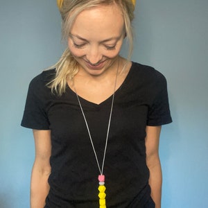 May include: A woman wearing a yellow headband and a black t-shirt is wearing a yellow, pink, and white silicone beaded lanyard with a silver clip.