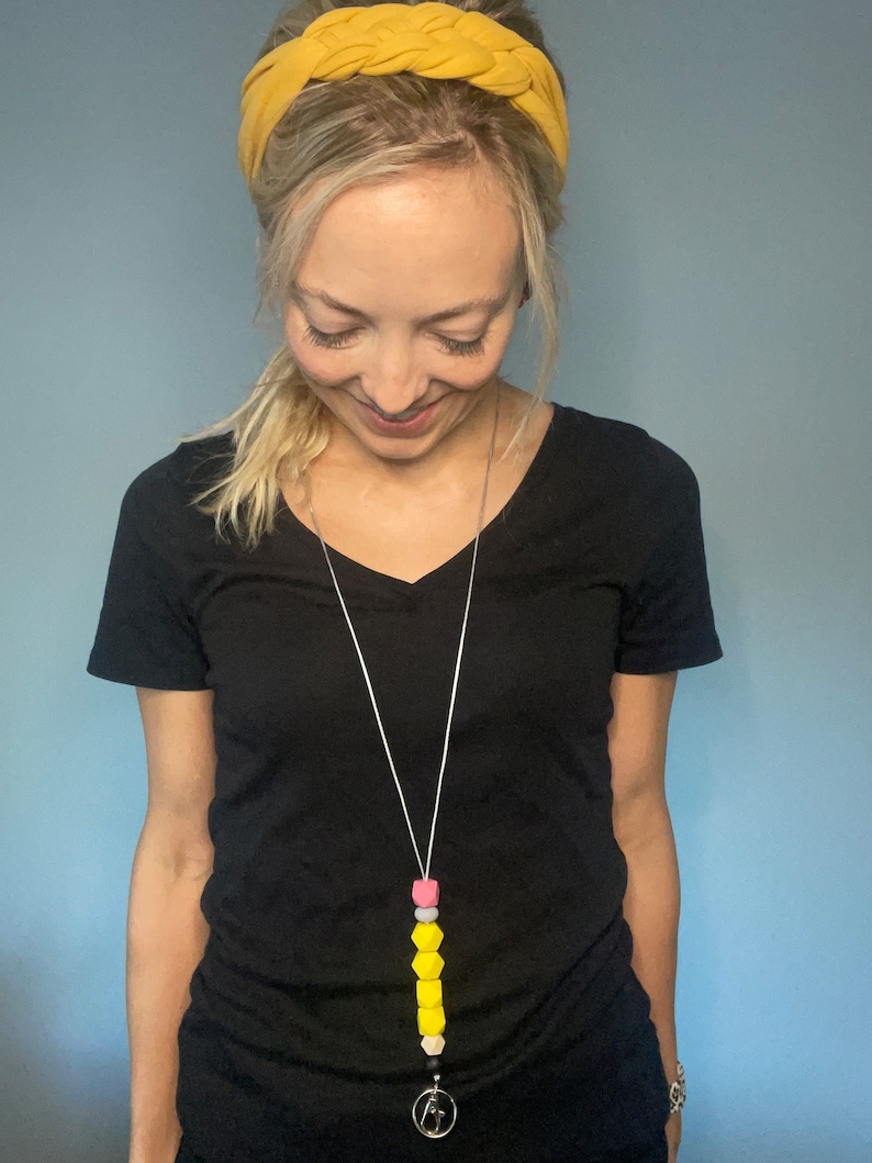 May include: A woman wearing a yellow headband and a black t-shirt is wearing a yellow, pink, and white silicone beaded lanyard with a silver clip.