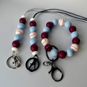 May include: A set of three beaded keychains with a baseball theme. The keychains are made with blue and burgundy silicone beads with white baseball designs. The keychains have silver, black, and silver lobster clasps.