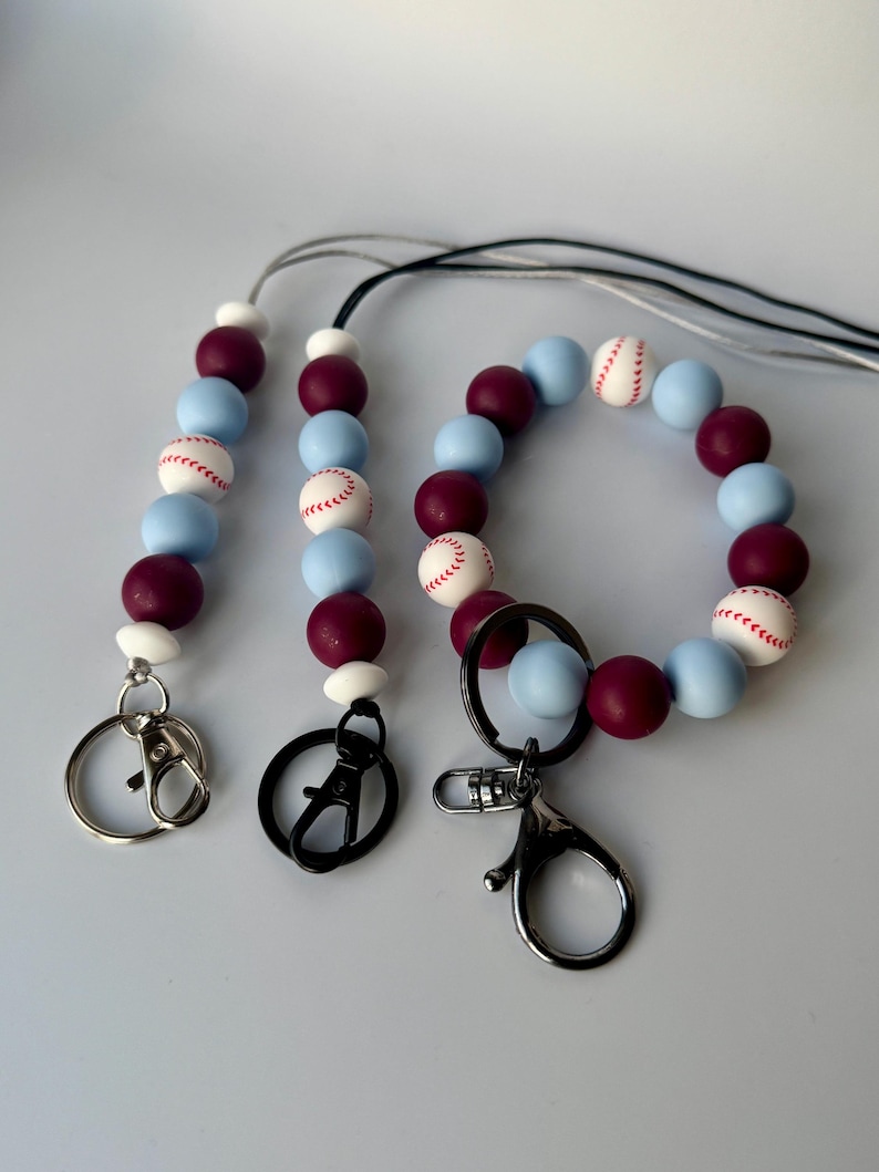 May include: A set of three beaded keychains with a baseball theme. The keychains are made with blue and burgundy silicone beads with white baseball designs. The keychains have silver, black, and silver lobster clasps.