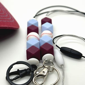 May include: A burgundy and light blue silicone beaded lanyard with baseball-themed charms. The lanyard has a black swivel clip and a silver lobster clasp.