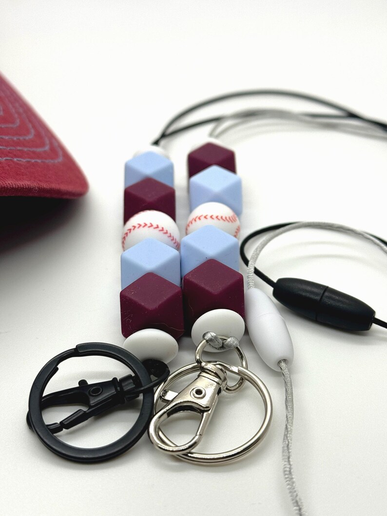 May include: A burgundy and light blue silicone beaded lanyard with baseball-themed charms. The lanyard has a black swivel clip and a silver lobster clasp.