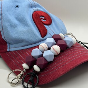 May include: A blue and red baseball cap with a white and red baseball charm attached to a silicone beaded lanyard with white and purple beads. The lanyard has two silver and black keychains attached.