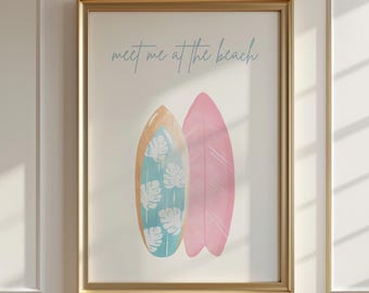 May include: A framed wall art print featuring two surfboards and the text "meet me at the beach" in blue script. One surfboard is light blue with white monstera leaf patterns, and the other is solid pink. The frame is a metallic gold color.