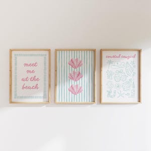 May include: Three framed wall art prints with light wood frames. The left print features the pink text "meet me at the beach" inside a blue striped border. The center print shows three pink seashells on blue stripes. The right print displays blue icons.