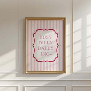 May include: A framed wall art print featuring a light pink and white vertical striped background. Centered is a white rectangular label with a dark pink scalloped border containing the text BUSY DILLY DALLY ING with a small heart icon.