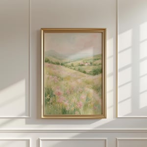 May include: A framed landscape painting featuring a soft-toned meadow filled with pink and green wildflowers in the foreground, a winding path leading toward a distant house, and rolling hills under a pale, hazy sky in a gold-colored frame.