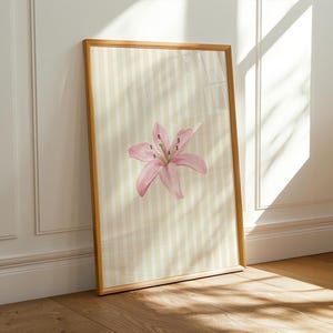 Puede incluir: Una lámina de arte rectangular en un marco de madera clara que presenta una flor de lirio rosa centrada sobre un fondo de rayas verticales de color crema y blanco. La obra se muestra en orientación vertical con un diseño sencillo.