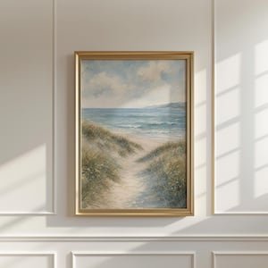 May include: A framed wall art print featuring a watercolor painting of a coastal landscape. The scene depicts a sandy path winding through green dunes toward a blue ocean under a cloudy sky, all enclosed within a metallic gold rectangular frame.