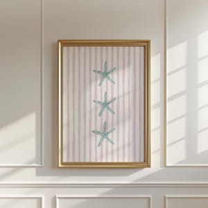 May include: A framed print with three pale blue starfish set against a pink and white striped backdrop. The artwork is in a gold-coloured frame, evoking a seaside theme. The framed print is displayed on a white wall.