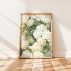 May include: A framed wall art print featuring a watercolor painting of white hydrangea flower clusters nestled among lush green leaves. The artwork is encased in a light-toned wooden frame, showcasing soft brushstrokes and botanical details.