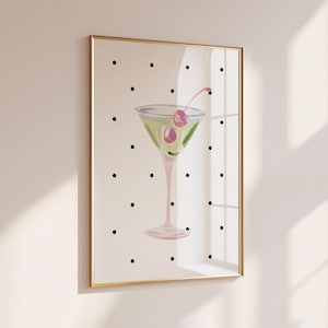 May include: A framed wall art print featuring a martini glass filled with a light green liquid and two pink cherries on a cream-colored background with a repeating pattern of small black polka dots. The frame is thin and metallic gold in color.