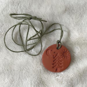 May include: A terracotta-colored pendant necklace with a nature-themed design. The round pendant features a tree, sun, moon, and stars. The necklace has a green cord. The pendant is approximately 1.5 inches in diameter.