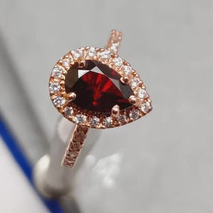 Peut inclure: Bague en or rose ornée d'une pierre précieuse rouge foncé en forme de poire, entourée de petits diamants clairs. La monture de la bague est également ornée de petits diamants clairs, créant un effet scintillant. Un beau bijou.