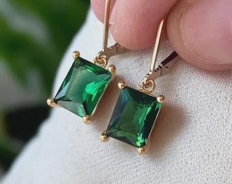 Pode incluir: Um par de brincos dourados com pedras preciosas verdes esmeralda com corte esmeralda. Os brincos têm um fecho de alavanca e pequenos detalhes dourados em cada canto das pedras retangulares.