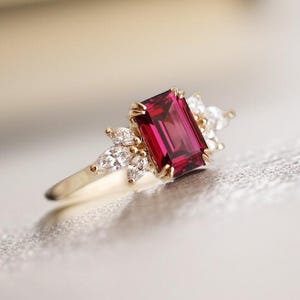 May include: A gold ring featuring a large, rectangular, deep red gemstone as the focal point. The ring is accented with smaller, clear gemstones clustered around the main stone, creating a sparkling effect. The band is a simple, polished gold.