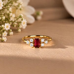 May include: A gold ring featuring a central emerald-cut red gemstone flanked by three small round clear stones on each side. The band is polished gold, and the setting holds the stones in a symmetrical arrangement, creating a refined appearance.