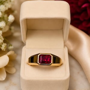 May include: A gold ring featuring a rectangular, faceted deep red gemstone set in a bezel mounting. The ring is displayed inside an open, light beige velvet-lined jewelry box, showcasing the polished metal band and the vibrant red stone.
