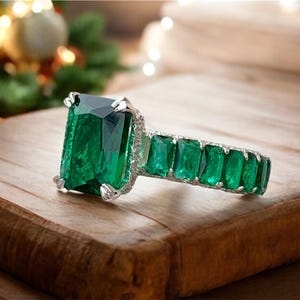 May include: A large emerald-cut green gemstone ring with a silver band. The ring features a large emerald-cut center stone and smaller emerald-cut stones along the band. The ring is displayed on a wooden surface.