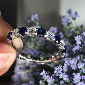 May include: A silver ring with alternating round, dark blue and clear gemstones. The ring is held up against a background of purple flowers.