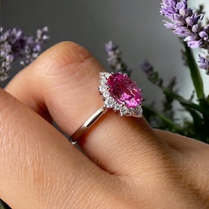 May include: A silver ring featuring a large, oval-cut pink gemstone surrounded by a halo of smaller, clear gemstones. The ring is set against a blurred background of purple flowers and a neutral backdrop.