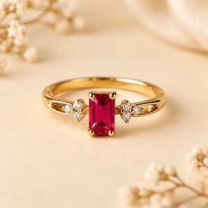 May include: A gold ring featuring a central emerald-cut red gemstone held by four prongs. The band is accented on each side by a cluster of three small, round, clear stones set in gold, creating a symmetrical design with a polished finish.