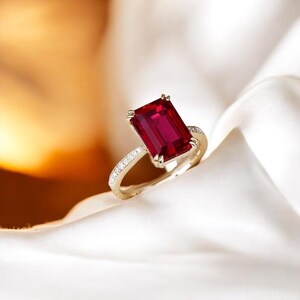 Puede incluir: Un anillo de oro con una gran gema rectangular de color rojo intenso. La banda de oro también presenta pequeñas piedras transparentes. El anillo se muestra sobre una tela blanca y sedosa.