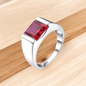 Può includere: Un anello d'argento con una gemma rettangolare rosso intenso. L'anello ha una finitura lucida e un design classico. La gemma è incastonata in una lunetta e la fascia è liscia e arrotondata. L'anello è esposto su una superficie di legno.