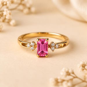 Può includere: Un anello in oro con una pietra preziosa rosa taglio smeraldo al centro, fissata da quattro griffe. La fascia presenta un gruppo di tre piccole pietre rotonde trasparenti su ciascun lato dell'incastonatura centrale, creando un design simmetrico.