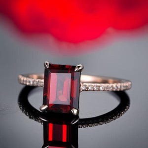 May include: A rose gold ring featuring a large, rectangular, deep red gemstone. The band is encrusted with small, clear stones. The ring is set against a blurred red and black background, creating a striking contrast.