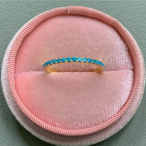 Puede incluir: Un anillo de oro con una banda de pequeñas piedras turquesas brillantes. El anillo se muestra dentro de un joyero de terciopelo rosa con un borde cosido rosa. El fondo es de color verde claro.