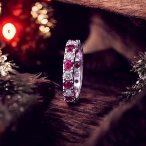 Könnte beinhalten: Ein silberner Ring mit abwechselnd roten Edelsteinen und klaren Diamanten. Der Ring liegt auf einer dunklen Holzoberfläche, mit verschwommenen Festtagsbeleuchtungen im Hintergrund. Der Ring ist ein Voll-Eternity-Band.