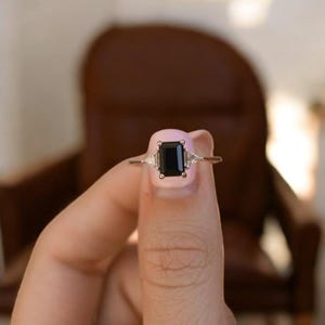 Könnte beinhalten: Ein goldener Ring mit einem großen, rechteckigen schwarzen Edelstein als Mittelpunkt, flankiert von zwei kleineren, dreieckigen weißen Edelsteinen. Der Ring wird vor einem unscharfen Hintergrund gehalten.