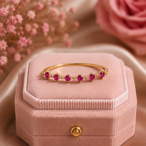 Peut inclure: Une fine bague en or ornée de six petites pierres précieuses rondes d'un rose vif disposées en ligne. L'anneau est délicat et poli, avec de minuscules perles d'or placées entre chacune des six pierres roses circulaires, créant un design symétrique.