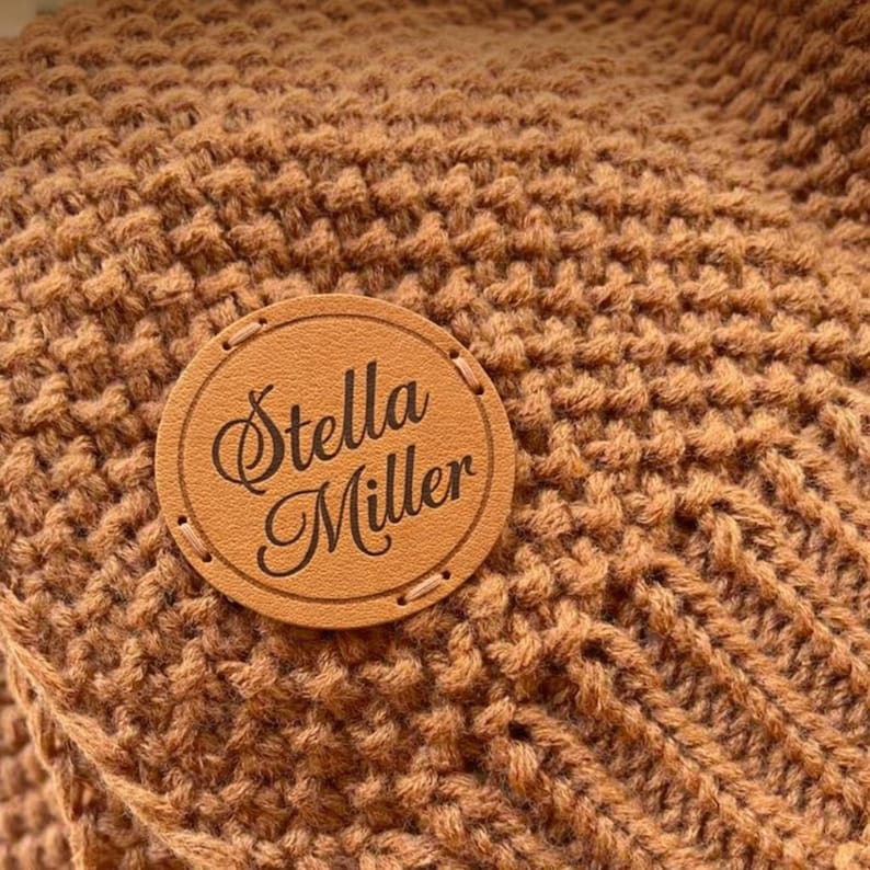 May include: Close-up of a textured, brown knitted fabric. A round, tan leather patch with the words "Stella Miller" in elegant script is stitched onto the fabric, adding a personalized touch to the textile.
