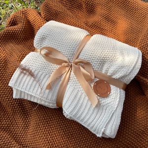 May include: A folded, off-white knitted blanket tied with a tan ribbon. The blanket is wrapped and presented on a textured, brown blanket. A small, round leather tag with the initials "D & T" is attached.