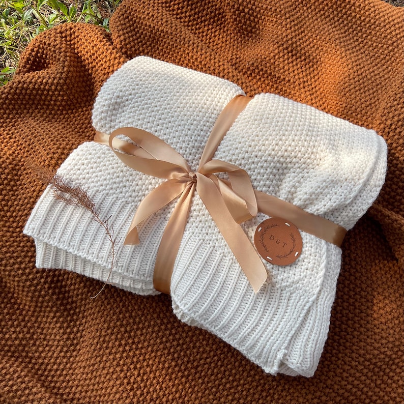 May include: A folded, off-white knitted blanket tied with a tan ribbon. The blanket is wrapped and presented on a textured, brown blanket. A small, round leather tag with the initials "D & T" is attached.