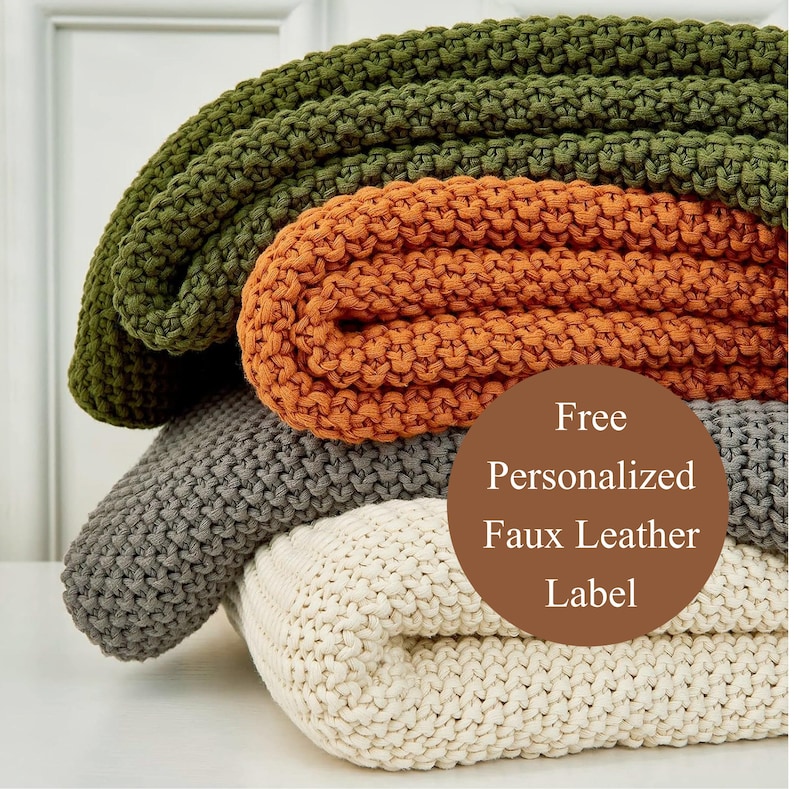 May include: A stack of four chunky knit blankets in olive green, orange, gray, and cream. The blankets are folded and stacked on top of each other. A brown circle with the text "Free Personalized Faux Leather Label" is in the image.