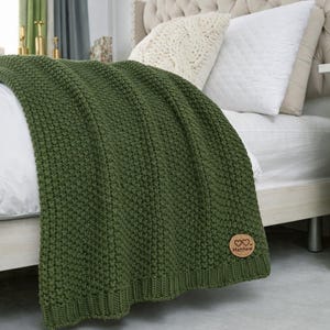 May include: A forest green knitted blanket draped over a bed. The blanket has a textured, raised pattern and a ribbed border. A small, round, wooden patch with two hearts and the name "Matthew" is attached to the blanket.