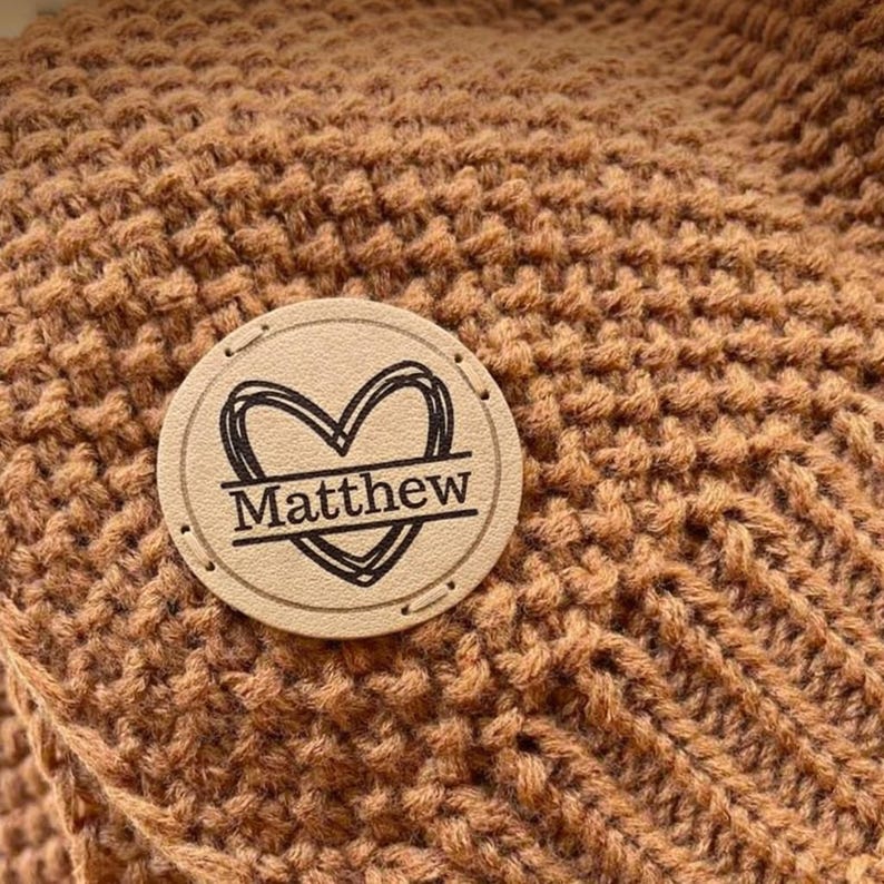 May include: Close-up of a brown knitted blanket with a circular leather patch. The patch features a heart design with the name "Matthew" in a black font. The blanket has a textured, woven pattern.