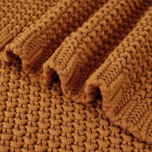 May include: Close-up of a folded, chunky knit blanket in a warm, golden-brown color. The thick yarn creates a textured surface with visible stitches and a soft, inviting appearance. The blanket's folds add depth and dimension.