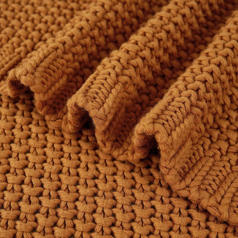 May include: Close-up of a folded, chunky knit blanket in a warm, golden-brown color. The thick yarn creates a textured surface with visible stitches and a soft, inviting appearance. The blanket's folds add depth and dimension.