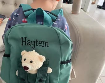 Könnte beinhalten: Ein tealfarbener Rucksack mit dem Namen "Haytem" in Schwarz gestickt. Der Rucksack hat einen Plüschteddybären in einer Tasche. Die Träger und Akzente sind teal. Das Kind trägt ein blaues Hemd mit Muster.