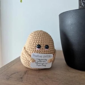 Puede incluir: Una figura de patata de ganchillo de color beige con ojos de botón negros. Sostiene un letrero que dice "Positive potato" y un mensaje motivador. La figura está colocada sobre una superficie de madera, con una planta y una maceta negra en el fondo.