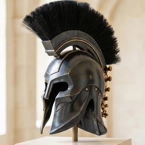 May include: A dark metallic Spartan-style helmet featuring a prominent black horsehair crest that runs from front to back. The helmet has a T-shaped face opening, curved cheek guards, and a series of small gold-toned rivets along the lower edge.