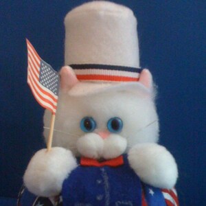 May include: A white plush cat wearing a white top hat with red, white, and blue stripes. The cat is holding a small American flag in its paw.