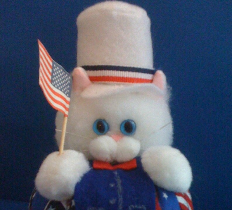 May include: A white plush cat wearing a white top hat with red, white, and blue stripes. The cat is holding a small American flag in its paw.