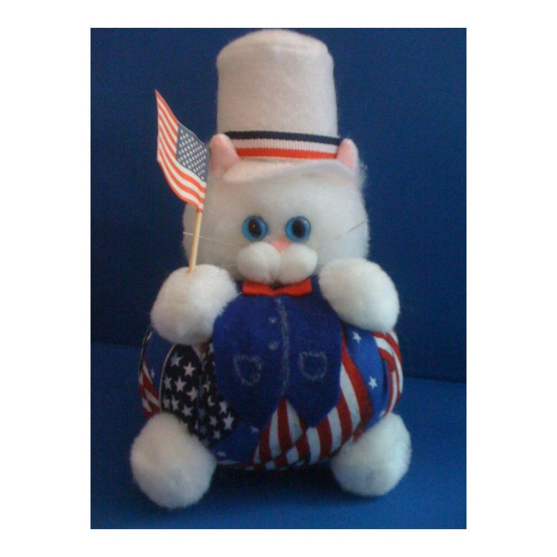 May include: A white plush cat wearing a red, white, and blue top hat and holding a small American flag. The cat is sitting on a round, fabric base with a patriotic design.