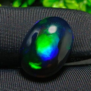 Könnte beinhalten: Ein polierter, oval geformter schwarzer Opal-Edelstein. Der Opal zeigt lebendige interne Farben von Grün und Blau mit einer hellen, reflektierenden Oberfläche. Der Edelstein liegt vor einem dunklen, strukturierten Hintergrund.